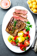Sliced medium rare grilled beef steak served on white plate with tomato salad and potatoes balls. Barbecue, bbq meat beef tenderloin. Top view, slate background, copy space