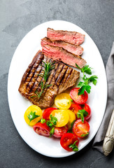 Sliced medium rare grilled beef steak served on white plate with tomato salad and potatoes balls. Barbecue, bbq meat beef tenderloin. Top view, slate background, copy space