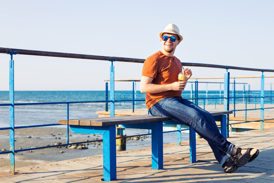 Portrait Of Cheerful Attractive Bearded Hipster Young Man Drinking Iced Coffee Or Frappe On The Background Of The Sea