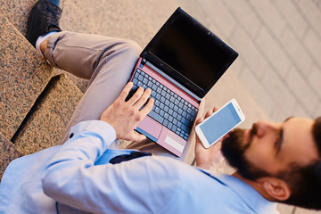 The stylish bearded male sits on a step and using laptop.