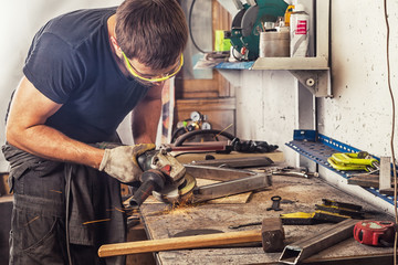 An adult man in work clothes and protective green glasses is engaged in manual labor and   grinder metal an angle grinder  on a wooden table in the workshop