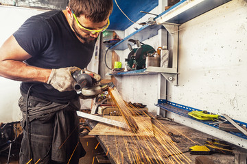 An adult man in work clothes and protective green glasses is engaged in manual labor and   grinder metal an angle grinder  on a wooden table in the factory, yellow sparks fly apart