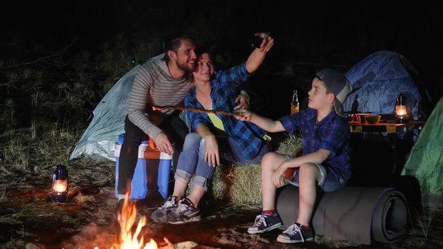 mother and father with son photographed on android in nature near bonfire, happy family makes photo on mobile phone near campfire in forest, parents with child into travel camp make selfi photo,