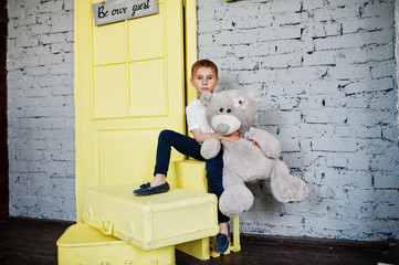Portrait of a little cute boy officially dressed posing with a teddy bear on yellow boxes in grey...