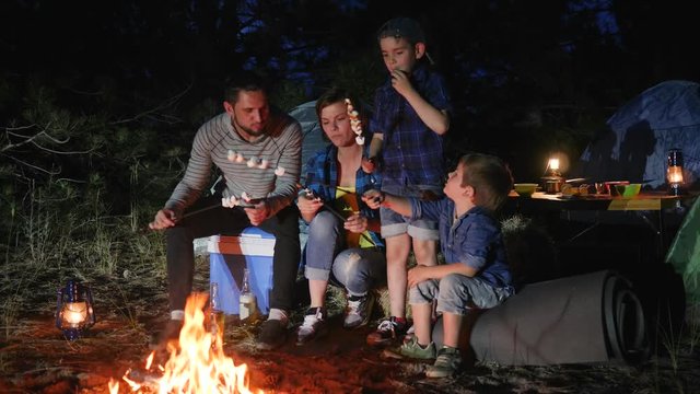 mom and daddy with kids resting to campground and roast zephyr on bonfire, parents with childs broil marshmallow on campfire to woodland, happy family fry marshmallow at fire in travel camp in forest,