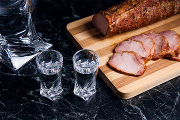 Bottle and shot glasses with vodka with slices of smoked meat on wooden cutting board.