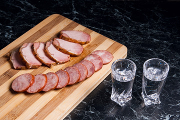 Shot glasses with vodka, slices of smoked meat and smoked sausage on wooden cutting board.