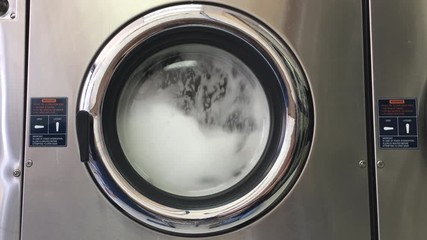 4K HD video of laundry agitating in a large industrial sized washing machine, stopping then re-starting. Bubbles forming viewable through door window.
