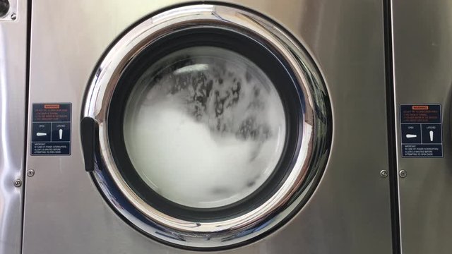 4K HD video of laundry agitating in a large industrial sized washing machine, stopping then re-starting. Bubbles forming viewable through door window.