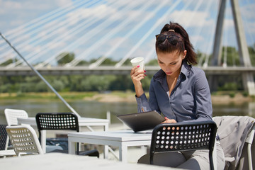 Beautiful young businesswoman with a disposable coffee cup, drinking coffee, and holding tablet in her hands against urban city background.