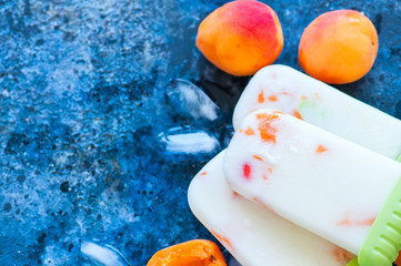 Homemade yogurt apricot popsicles on blue background with ice and ripe fruits. Healthy summer food concept with copy space.