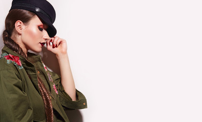 Beautiful young girl with bright make-up in a green trench coat and a black cap stands in the studio on a white background. Military uniform, army, fashion, beauty, hairstyle - fish tail. clothing.