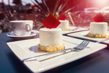 A set of brownies with red chocolate on white plates and a cup of coffee on a table in a street cafe. Sunset.