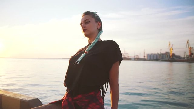 Young stylish funky girl with green hair riding roller skates and dancing near sea port during sunset, slow motion