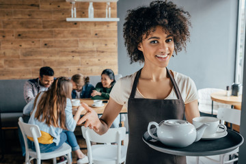 Smiling african american waitress holding tray with tea and customers sitting behind her in cafe