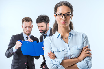 portrait of businesswoman in eyeglasses with arms crossed looking at camera while colleagues discussing work behind