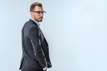 back view of confident businessman in eyeglasses and formal wear looking at camera isolated on blue
