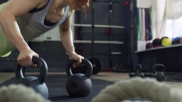 PAN of muscular female athlete with strong arms using kettlebells as bars and doing push-ups in gym