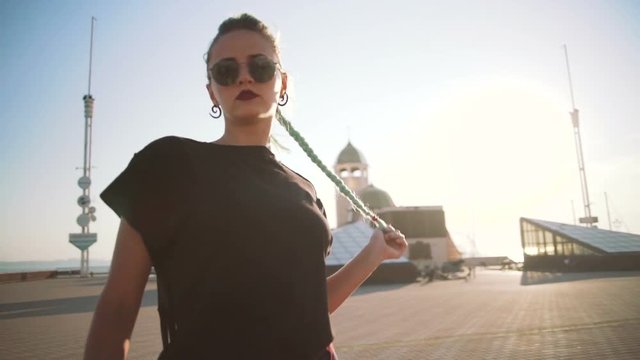 Young stylish funky girl with green hair riding roller skates and dancing near sea port during sunset, slow motion
