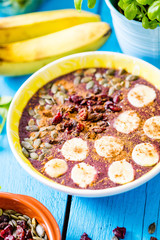 Close Up View of Smoothie Bowl with Blueberry, Banana, Pumpkin Seeds, Cinnamon and Cranberries on Blue Wooden Table, Vertical View