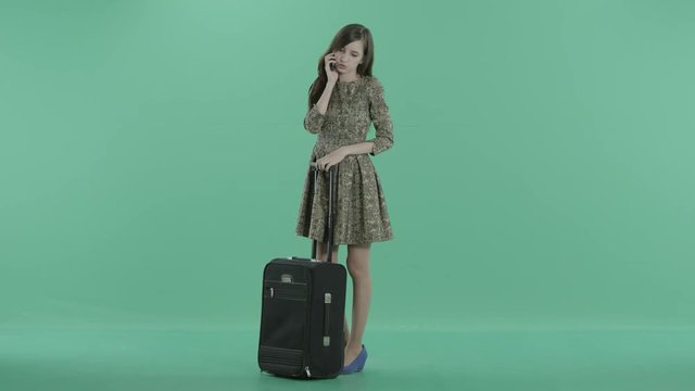 a girl in dress is standing with suitcase and talking on the phone