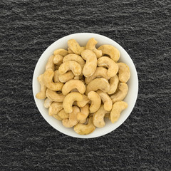 Top view of raw cashews in a small white bowl on a black slate table.
