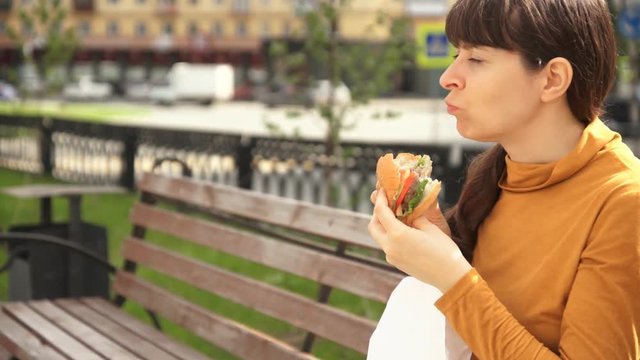 Close-up shot of a young brunette woman in a yellow turtleneck eating a hamburger sitting on a wooden bench outdoors next to the road. Lunch break on a summer or autumn day.