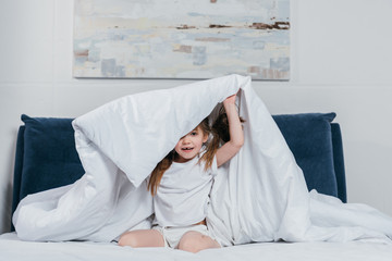 portrait of little caucasian girl hiding under blanket in bedroom