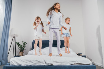 multiethnic little girls in pajamas jumping on bed at home