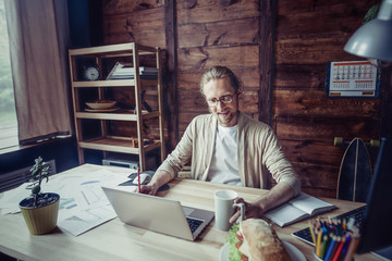 Freelancer man sitting at table holding cup of tea. Hipster man sitting working with laptop drinking tea.