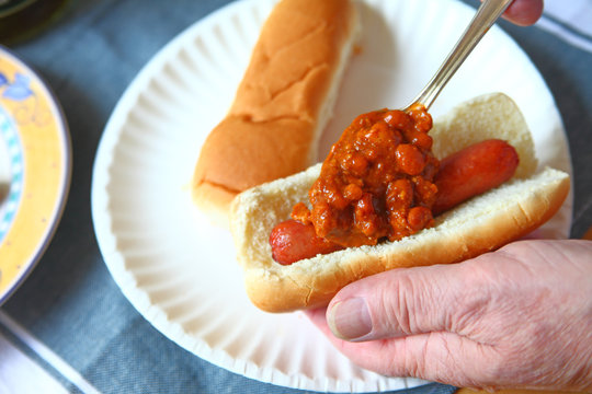 A Man Spoons Chili With Beans Onto A Hot Dog Over A Paper Plate