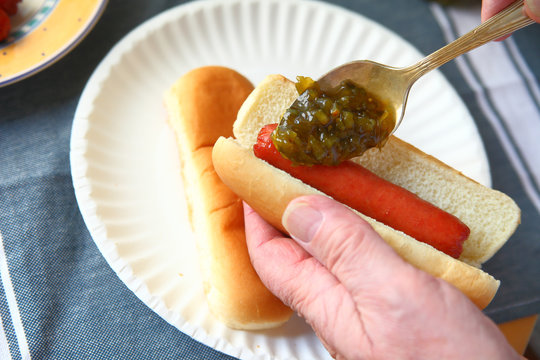 A man adds sweet pickle relish to a hot dog over a paper plate with additional bun
