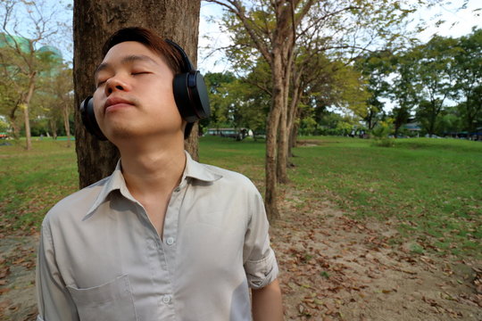Happy young man listening to music with headphones and leaning a tree in the public outdoor park.