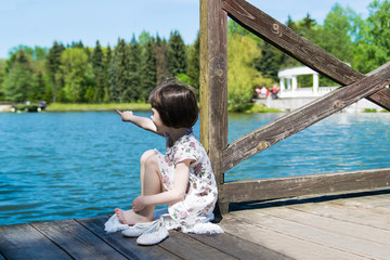 A little young girl is sitting on a wooden pier. Summer day