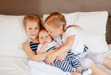 Lifestyle portrait of cute white Caucasian girls sisters holding kissing little baby, sitting on bed indoors. Older siblings with younger brother sister newborn. Family love bonding together concept.