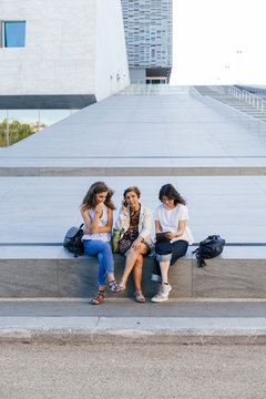 Mother and two daughters sitting near a modern building use their smart phone and tablet