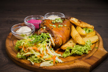 Fried pork leg drumstick, knee of a wild boar with vegetables of red and yellow tomatoes, garlic, parsley and basil and potatoes on a wooden background