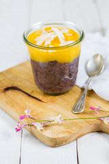 
Black rice pudding with mango and coconut flakes in the glass jar.  White background, wooden table.