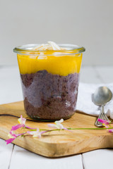 
Black rice pudding with mango and coconut flakes in the glass jar.  White background, wooden table.