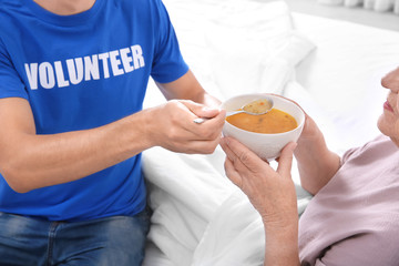 Obraz na płótnie Canvas Young male volunteer feeding elderly woman in light room