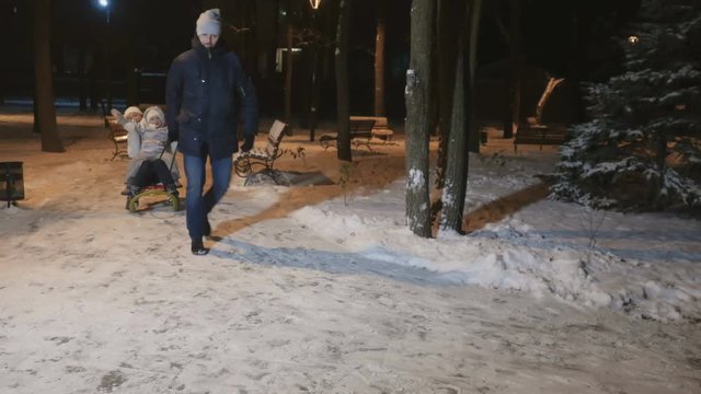 Father rolls a wife with little daughter on the sled in the evening winter park