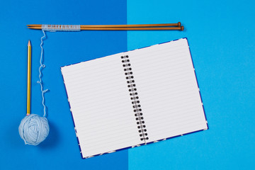 Knitting needles, blue yarn ball and open paper notebook on blue background