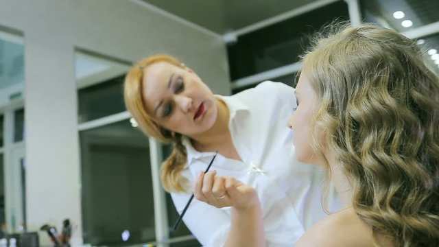 Professional makeup artist applying eye pencil and eye brush to blond models eyelid for doing eyelid arrows. Young attractive girl with long curly blond hair and blue eyes.