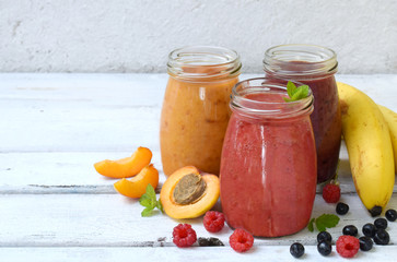 Orange, red and purple healthy smoothies of fresh ingredients - bananas, apricots, raspberries and blueberries on a light background. Copy space