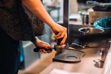 Close up shot of female barista carefully tamping fresh arabica coffee on top of bar, ready to extract powerful espresso shot out of machine, in cool downtown cafe