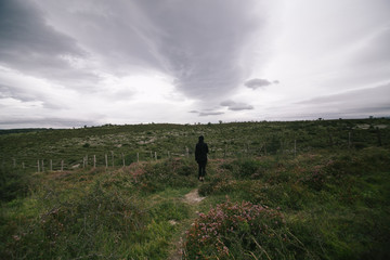 Fototapeta premium Silhouette of lonely traveler in black outfit standing in middle of field or meadow on grey rainy day with sad atmosphere of loneliness and doom