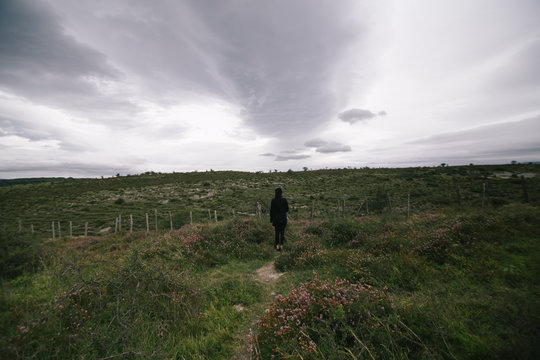 Silhouette of lonely traveler in black outfit standing in middle of field or meadow on grey rainy day with sad atmosphere of loneliness and doom