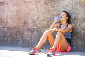 Fototapeta na wymiar Young smiling female resting after an active training with water bottle while siting on sidewalk, satisfied fit woman resting after an active training, drinking cold water.