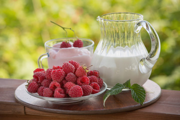 A jug of milk, a bowl full of raspberries and raspberry yogurt on a wooden stand. Red berries and drinks on the green natural background.