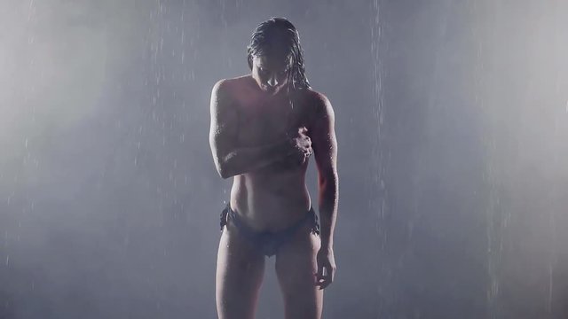 girl bodybuilder standing in the fog in the rain in a dark room. she's almost naked covering her breast with hand. average plan